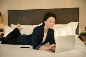 Beautiful young Caucasian business woman lying down on a bed in a hotel bedchamber and working on laptop. Distant work, remote, online concept photo