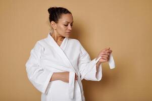 Attractive woman tying a belt on a white waffle robe posing against a beige background with copy space. Skin care and spa concept photo