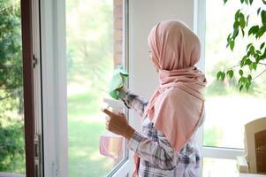 Arab Muslim woman with a covered head in a hijab washes windows, removes stains, sprays detergent and wipes with a rag enjoying a general Spring cleaning of the house photo