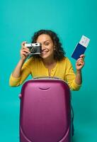 Cheerful smiling Hispanic woman holding a passport with ticket and boarding pass and a vintage camera, photographing, smiling toothy smile looking at camera against blue colored background. Copy space photo