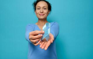 Focus on blue satin ribbon, symbolic bow color of World Diabetes Awareness Day, in the hands of beautiful mixed race woman wearing a blue sweatshirt, isolated on colored background with copy space photo