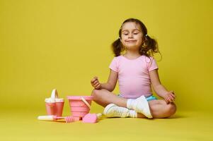 Adorable little girl meditating with closed eyes, sitting near set of beach toys on yellow background with copy space photo