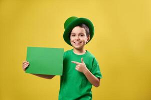 Happy boy in green clothes and Irish hat puts his index finger to a sheet of paper with copy space and smiles while posing on a yellow background. Saint Patrick's Day photo