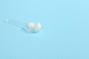 Pure refined white sugar cubes on a transparent measuring spoon isolated over colored blue background with copy space for medical advertising. World diabetes day awareness concept, 14 November. photo