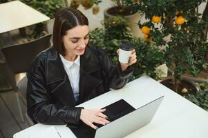 Overhead view of beautiful business woman, copywriter, developer, freelancer working remotely on laptop, writing text, enjoying coffee break in outside summer cafeteria with tangerine decorative tree photo