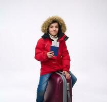 Cheerful preteen, elementary aged boy in bright red down jacket sitting on his luggage, looking at camera and showing passport with boarding pass. Winter holidays, travel concept with copy ad space photo