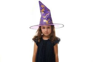 retrato de maravilloso confidente pequeño niña vistiendo un mago sombrero y vestido en elegante carnaval vestido, mirando a cámara posando con cruzado brazos en contra blanco fondo, Copiar espacio. foto