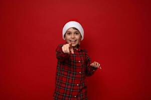 Adorable preadolescent boy in red checkered shirt wearing a Santa hat points directly to camera, smiles with beautiful toothy smile posing against colored background with copy space for Christmas ad photo