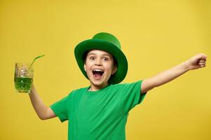 Happy boy in green T-shirt and leprechaun hat with hands up holds a glass with green drink and express happiness celebrating the Saint Patrick's Day. Copy space photo