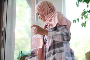 Middle aged beautiful Muslim woman with her head covered in pink hijab washing windows during spring cleaning in the house. Housekeeping, chores, cleaning concept photo