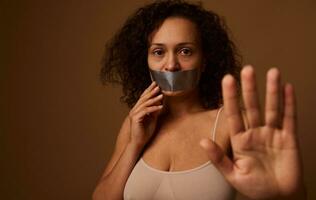 Desperately scared crying woman with tears in her eyes, sealed mouth shows STOP sign with hand looks at camera, isolated on dark beige background. Social concept of elimination violence against women photo