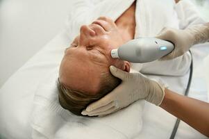 Close-up of beautician hand holding modern ultrasonic medical apparatus and performing face lifting and rejuvenating anti-aging beauty treatment on face of a relaxed mature man in wellness spa clinic photo