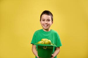 sonriente chico sostiene un duende verde irlandesa sombrero lleno de manzanas y sonrisas a cámara. Santo patrick's día. amarillo antecedentes foto