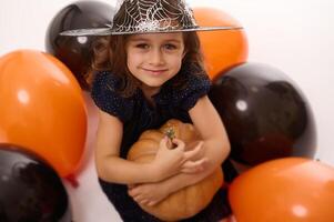 4 4 años antiguo pequeño niña en bruja disfraz y mago sombrero obras de teatro con globos y un calabaza aislado en blanco antecedentes. concepto de un niño teniendo divertido a Víspera de Todos los Santos fiesta foto