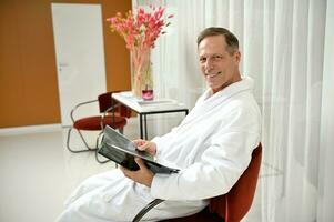 Handsome mature European man dressed in white terry bathrobe, smiles looking at camera while relaxing in private room of a wellness spa centre, sitting on an armchair with a magazine in his hands photo