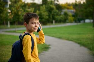 Side view of a schoolboy wearing yellow sweatshirt with backpack walking on the path in public park, going home after school, talking on mobile phone, smiling with toothy smile to the camera photo