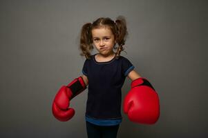 Cute European 4 years old baby girl wearing red boxing gloves poses against gray wall background with copy space for advertisement for Boxing Day photo