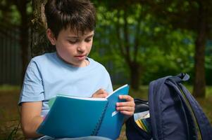 Close-up of Hispanic smart preteen school child boy writing on copybook, doing homework outdoors. Back to school concept photo