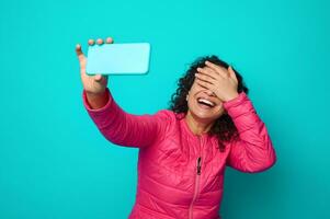 Attractive dark-haired awesome woman with curly hairstyle, wearing pink jacket, covering her eyes, smiles with beautiful toothy smile with making selfie on her smartphone. Blue background Copy space photo