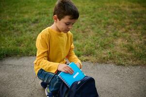 Adorable elementary aged schoolboy sits on his knees on a path in the park, puts a notebook and pencil case in a backpack, returning home after shooting. Back to school concept photo