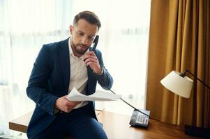 Busy puzzled handsome middle aged European man, entrepreneur, successful businessman holding a stack of documents and negotiating while talking on landline phone in hotel room during business trip photo