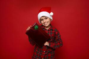 Handsome preadolescent boy in Santa Claus hat and red plaid shirt poses over colored background with Christmas gift, smiled with beautiful toothy smile looking at camera. Copy space for advertisement photo