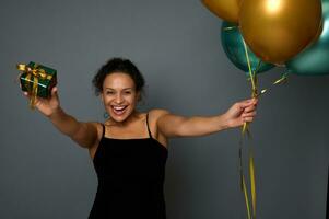 Cheerful African woman in black dress celebrates birthday party, holds a luxury gift and shiny air balloons in outstretched hands, smiles looking at camera isolated on gray background with copy space photo