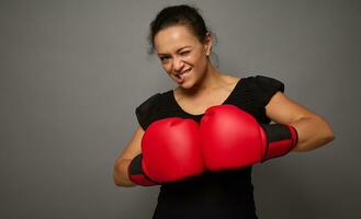 Strong concentrated African woman looks at camera posing against gray wall background with red boxing gloves. Concept of Black Friday and boxing Day, Blow to prices with copy space for advertisement photo