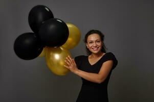 Cheerful African woman with beautiful smile poses with inflated black and gold air balloons against gray wall background with copy space. Concept of Black Friday and gold credit shopping card photo