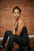 retrato de un hermosa deportivo mujer en negro ropa de deporte sentado en un medicina aptitud pelota un posando mirando a cámara en contra rojo ladrillos pared antecedentes de un gimnasio clase foto