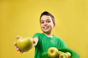 Cute little smiling caucasian boy holds an Irish leprechaun green hat full of apples in his hands and shows a green apple to camera. Saint Patrick's Day. Copy space photo