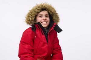 Waist length portrait of handsome cheerful preadolescent, school aged boy wearing bright red down jacket with hood, smiling toothy smile looking at camera isolated on white background with copy space photo