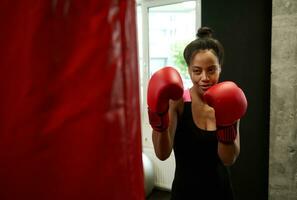 Beautiful African fit woman with perfect physique posing at camera wearing red boxing gloves, hitting punching bag in gym. Female boxer training hard during martial combat art photo