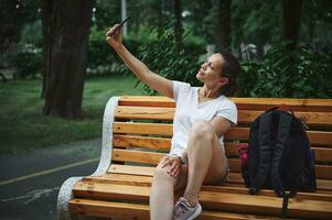 Young multi-ethnic pretty woman using smartphone, taking a selfie, sitting on a wooden bench while resting in the park photo