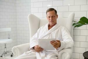 Handsome mature European man dressed in white bathrobe, with a blank paper sheet, sitting armchair in luxurious wellness spa centre photo