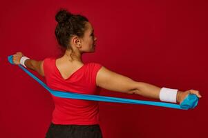 Close-up of beautiful Hispanic fit sportswoman working out with elastic rubber strap against red colored background with copy ad space photo