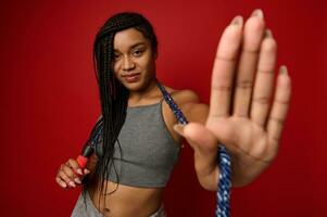 Fashionable sporty cheerful African woman with stylish dreadlocks posing against a red background with a jumping rope around her neck, gesturing, showing a STOP sign, looking at the camera photo