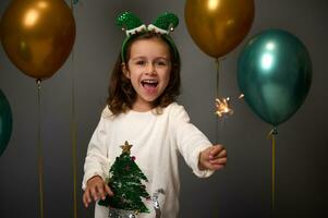 Adorable beautiful child, cute baby girl having fun lightening the Christmas sparklers, laughing looking at camera against gold and green metallic air balloons on gray wall background with copy space photo