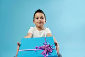 Cute little boy holds a blue gift with pink bow out to the camera while sitting on a blue background. Celebration Birthday concept photo