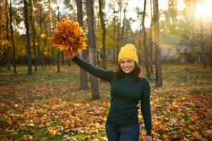 Adorable young woman raising hands with collected dry bouquet of fallen autumnal maple leaves and cutely smiling with beautiful toothy smile posing against autumn park with falling sun rays at sunset photo