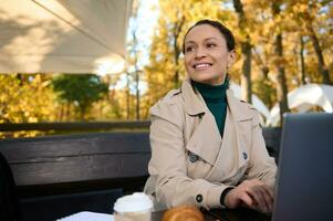 encantador negocio mujer en zanja Saco sentado a el mesa de un de madera café en el roble arboleda y mirando el lado, sonriente con con dientes sonrisa mientras trabajando en ordenador portátil en hermosa otoño antecedentes foto