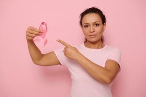 Portrait of serene Middle Eastern ethnicity woman in pink T-shirt pointing to a satin ribbon in her hand, looking at camera, isolated on colored background with copy space. Breast Cancer Day concept photo