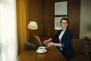 Young businesswoman, entrepreneur, successful sales manager in eyewear and business casual suit sits at a desk and works on laptop in hotel room during business trip photo