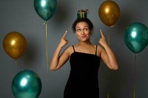 africano bonito mujer en noche vestir puntos dedos en un Navidad regalo en su cabeza, mira arriba, poses en contra gris pared antecedentes decoración con dorado verde metálico aire pelotas. celebracion concepto foto