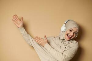 maravilloso musulmán mujer en hijab escuchando a el música en auriculares, señalando manos en beige antecedentes con Copiar espacio foto