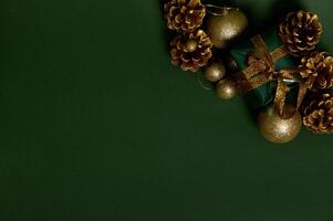 Flat lay of a present in shiny wrapping gift paper with golden bow, pines cones and gold Christmas tree toys laid out on the corner of green background. Copy space for advertisement photo