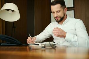 Attractive handsome successful confident caucasian businessman, journalist, businessman on business trip, writes and makes notes in notebook and drinks coffee, sitting at a table in a hotel apartment photo