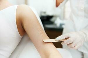 Close-up. The beautician holds a wooden spatula and applies a transparent gel with anesthetic to the woman's hand and prepares the skin for the laser hair removal procedure. photo