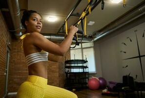 Side view of an attractive African woman doing glute exercises, performing side lunges while working out with suspension straps in fitness studio. Fitness, stretching, active lifestyle concept photo