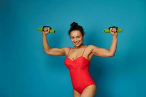 Athletic woman in red swimsuit posing with dumbbells, straining biceps muscles on blue background photo